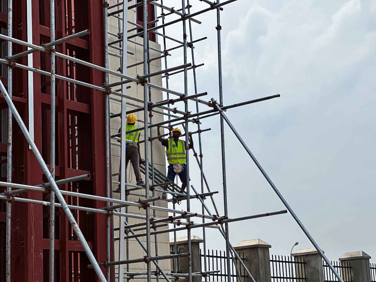Two workers in safety vests and helmets work on scaffolding, set against a cloudy sky.
